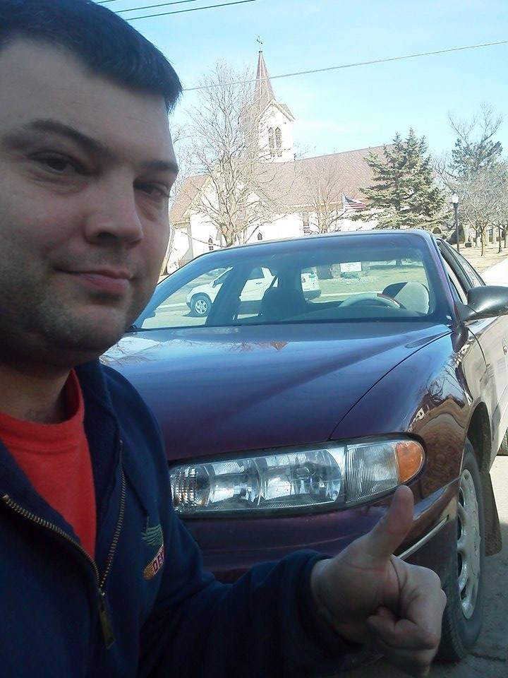 Scott Lawrence giving a thumbs-up next to his maroon Buick Century, with a white church in the background
