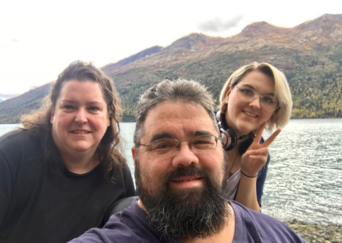 Eric with family, a selfie in front of a lake with mountains behind