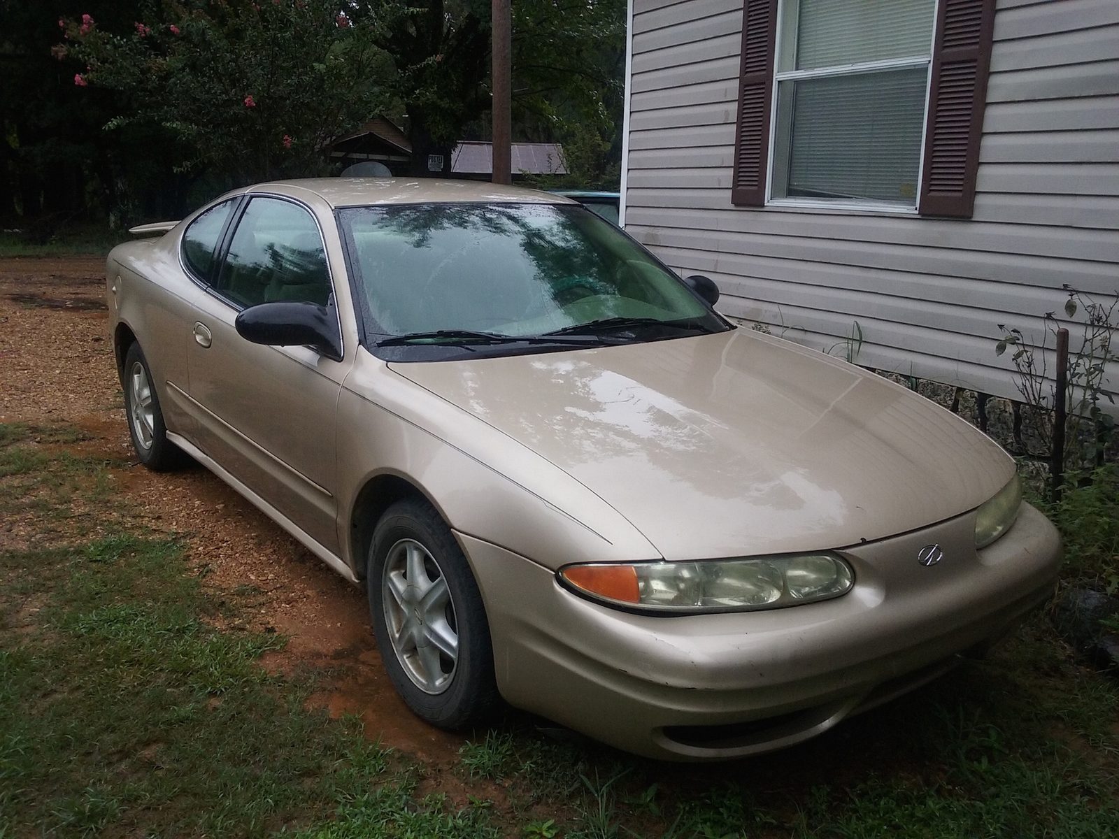 Gold Oldsmobile Alero coupe parked on grass beside a house